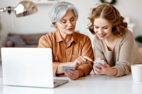 Young woman teaching grandmother to do online shopping Young granddaughter smiling and showing smartphone to elderly lady with credit card while shopping online at home together